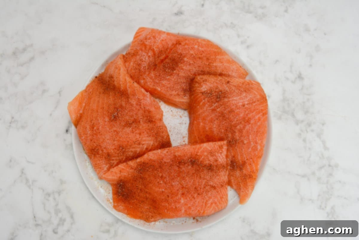 Raw, marinated salmon fillets now richly coated with an aromatic blend of dry spices, including salt, pepper, paprika, and chipotle powder, arranged on a white plate.
