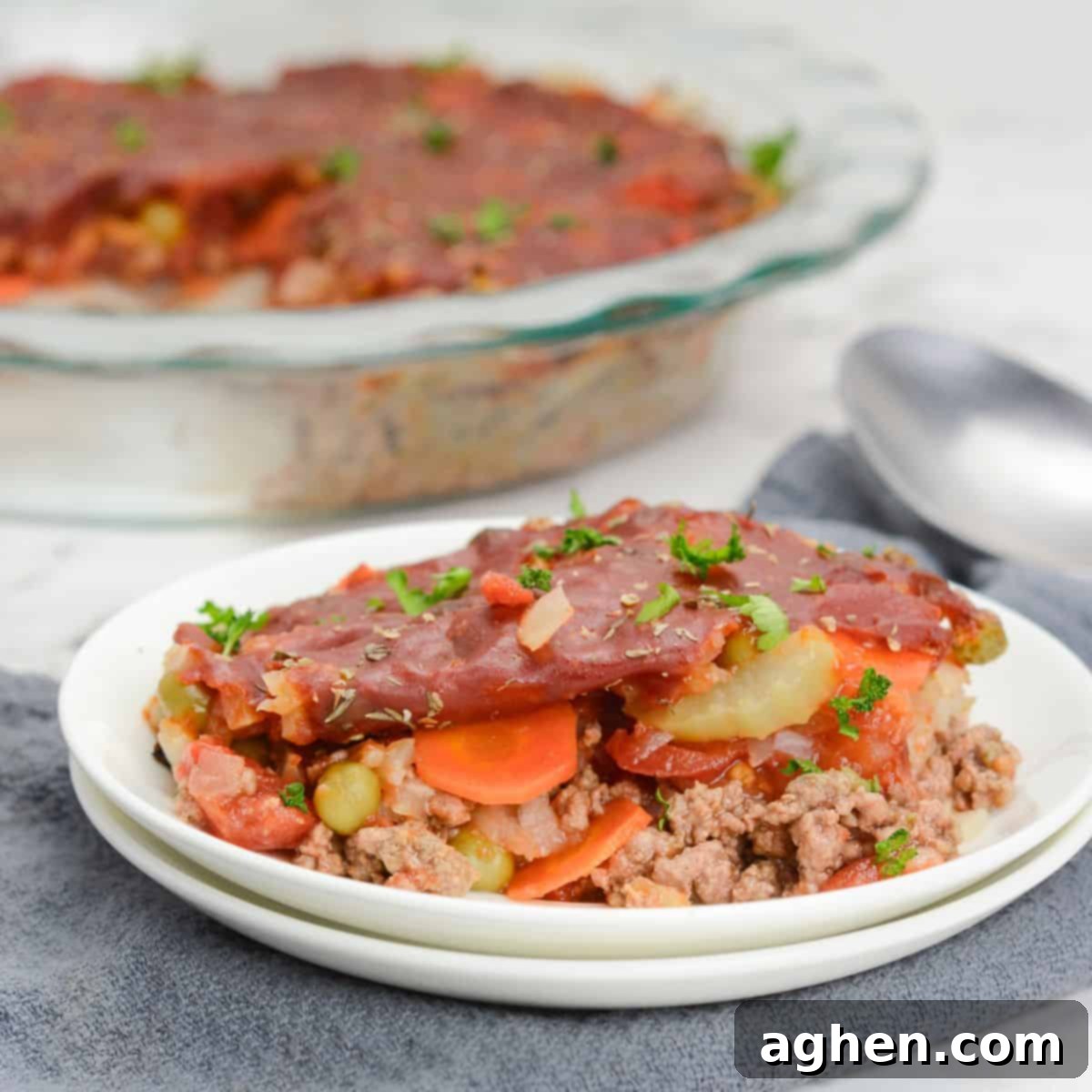 A perfectly portioned plate of Shipwreck casserole, topped with fresh herbs, is artfully placed on a gray napkin. A serving spoon and the full casserole dish are blurred in the background, creating an inviting culinary scene.