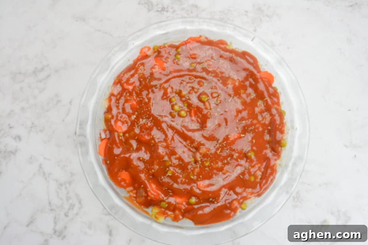 A pie plate shows the fully assembled Shipwreck casserole, beautifully topped with a smooth, red layer of condensed tomato soup and a final sprinkle of seasoning, ready for baking.