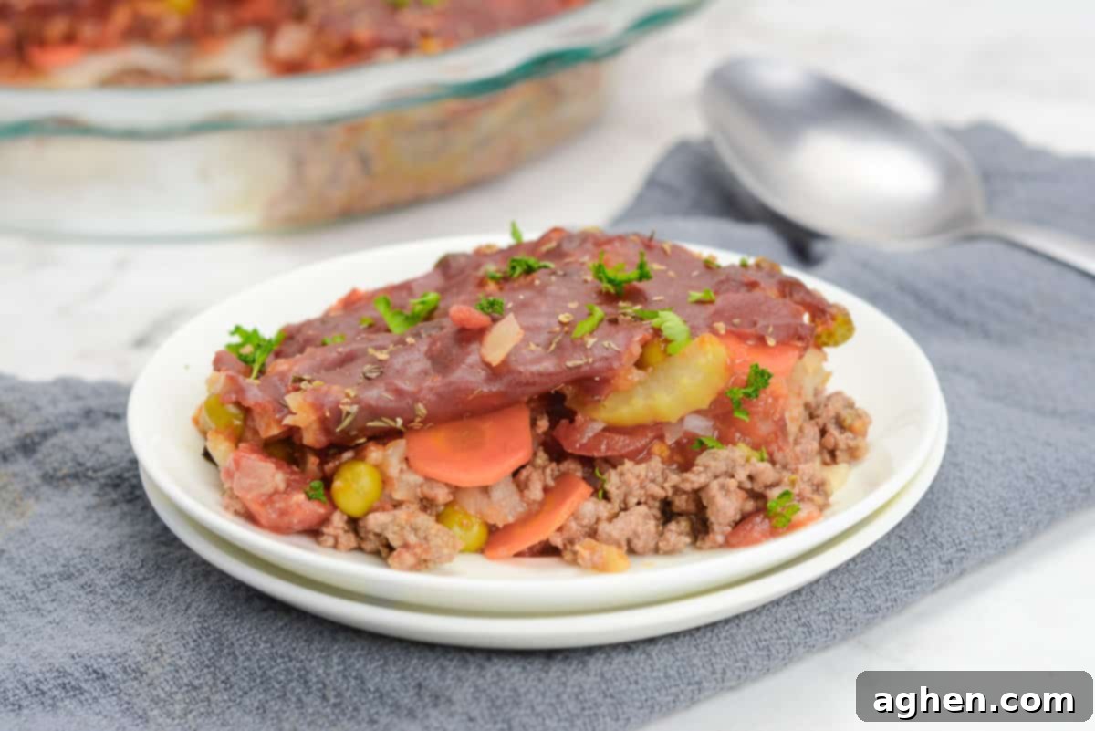 A generous plate of Shipwreck casserole, freshly baked and steaming, is presented on a textured gray napkin, with a serving spoon and the full casserole dish visible in the soft-focus background.