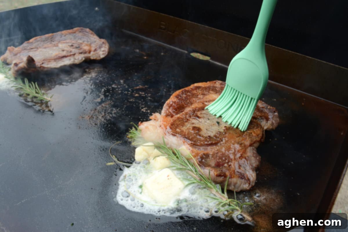 From Novice to Griddle Guru: 10 Hard-Earned Lessons After 3 Years on the Blackstone 3 Two sizzling steaks cooking on a Blackstone griddle next to melting butter and fresh rosemary sprigs. A blue silicone brush bastes one steak with butter, enhancing its flavor.