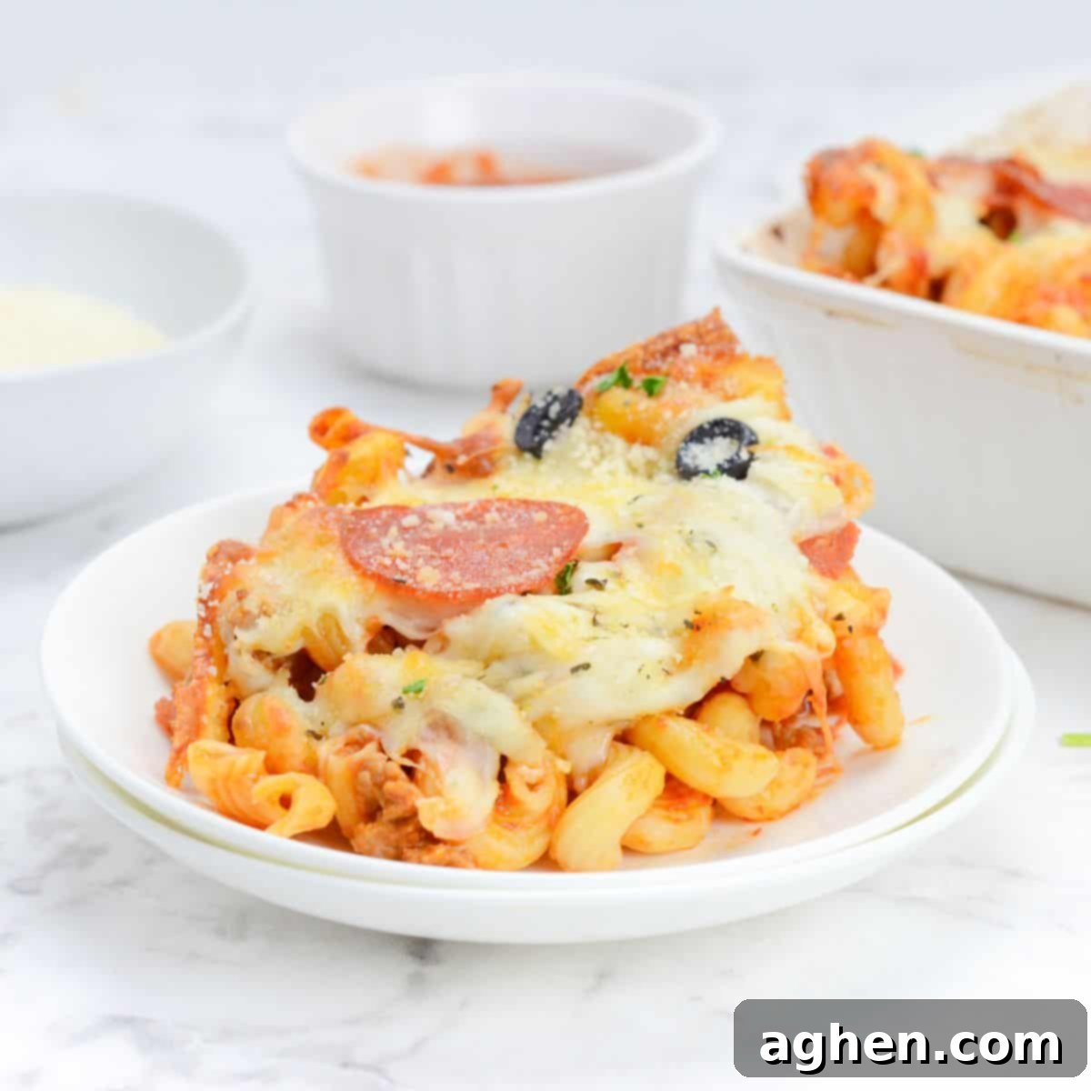 Single serving of pizza casserole is scooped onto a plate. In the background there is a casserole dish and two small bowls with sauce and parmesan.