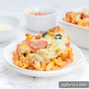 Plate of pizza pasta sits on white background. A bowl of parmesan, bowl of marinara and the casserole sit behind.