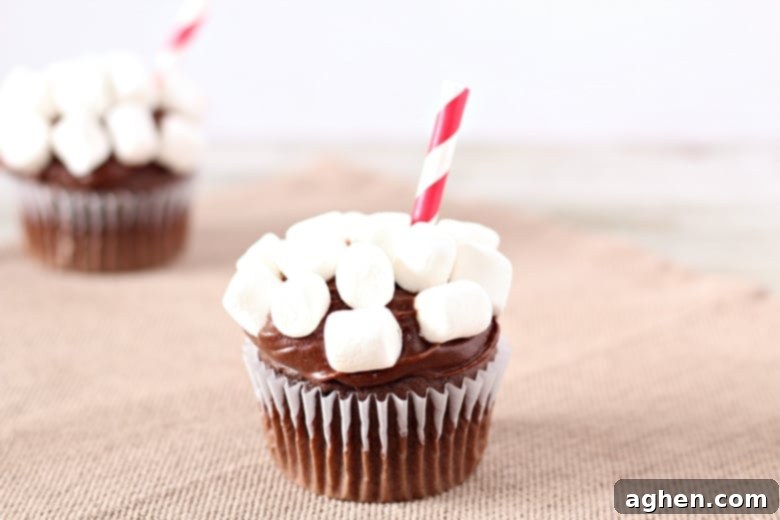 Close-up of a finished hot chocolate cupcake, with a striped straw inserted and mini marshmallows on top of chocolate icing.