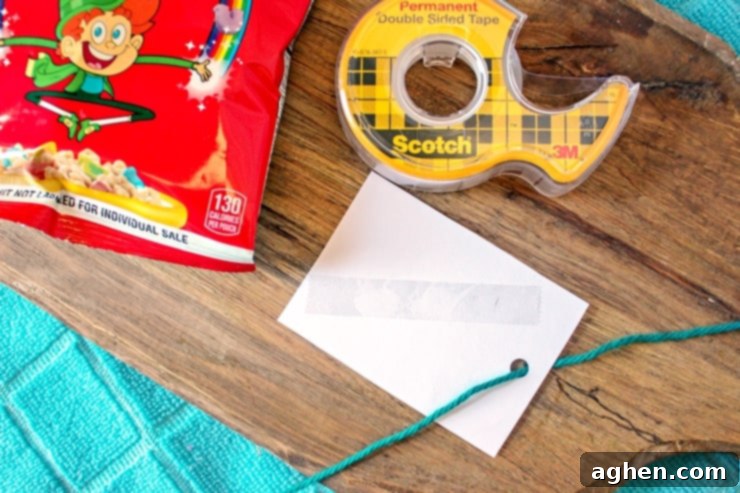 A gift tag turned over with tape on the back and yarn through the hole on top. Roll of double sided tape and a bag of Lucky Charms can be seen next to the tag.