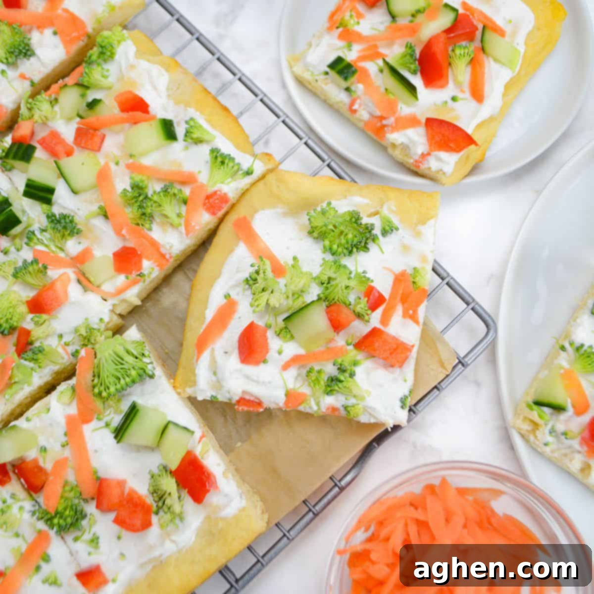 Veggie pizza cut into slices sits on top of a cooling rack lined with parchment. Two small plates with slices surround it and a small bowl of shredded carrots.