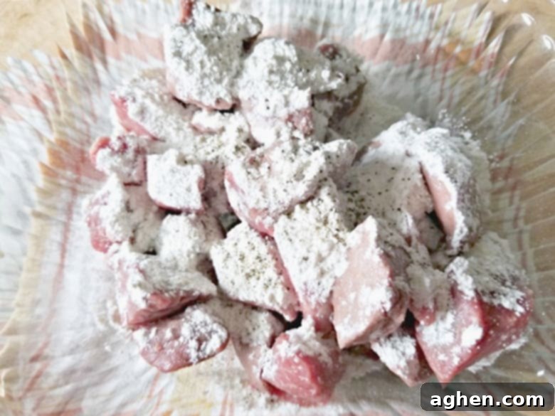 Close-up shot of cubed beef being tossed with olive oil, flour, salt, and pepper in a mixing bowl, ready for browning.