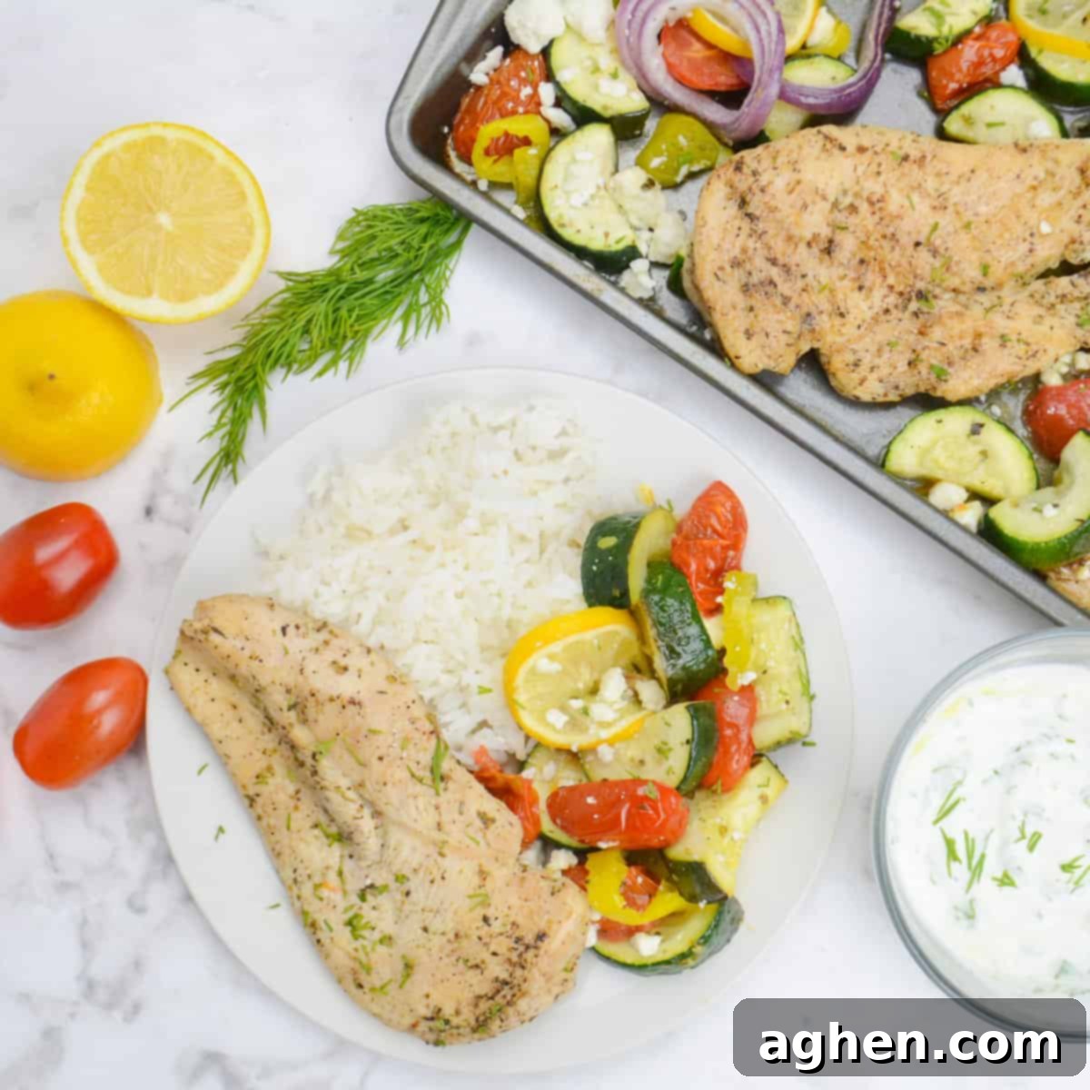 Effortless Delicious Weight Watchers Greek Sheet Pan Chicken 10 A white plate with a serving of chicken, roasted vegetables and white rice.To the side sits the sheet pan with additional servings, a bowl of tzatziki, a cut lemon, dill and tomatoes.