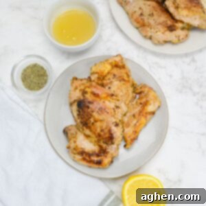 Small plate with two cooked chicken thighs. To the side sit a small bowl of honey, a bowl of oregano, a sliced lemon, a white napkin and off to the side a plate of chicken.