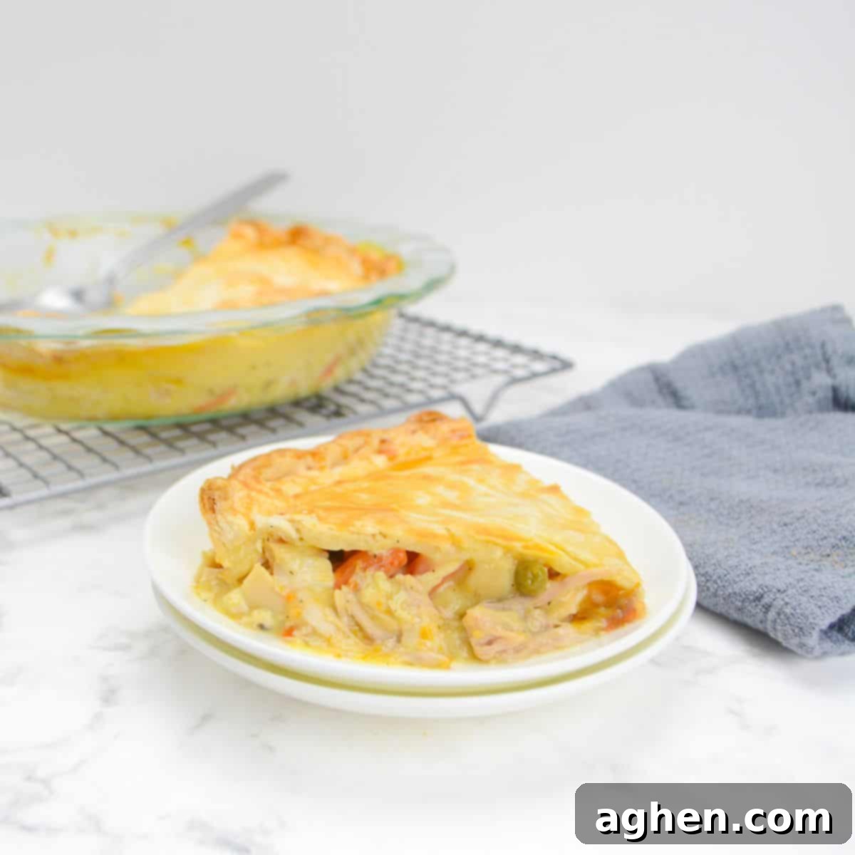 A single slice of golden-brown Weight Watchers Chicken Pot Pie rests on a white plate beside a grey napkin. In the background, the casserole dish sits on a cooling rack with a serving spoon.