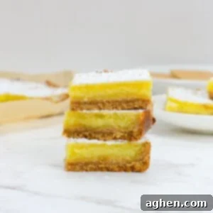Three sliced and stacked lemon bars with powdered sugar. In the background sit a plate of lemon bars and parchment with sliced bars.