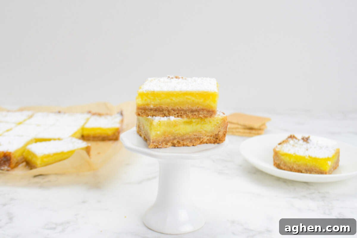 A small single cupcake stand with two sliced lemon bars placed on top. In the background are a plate with one lemon bar, a stack of graham crackers and a sheet of parchment with the remaining sliced bars.