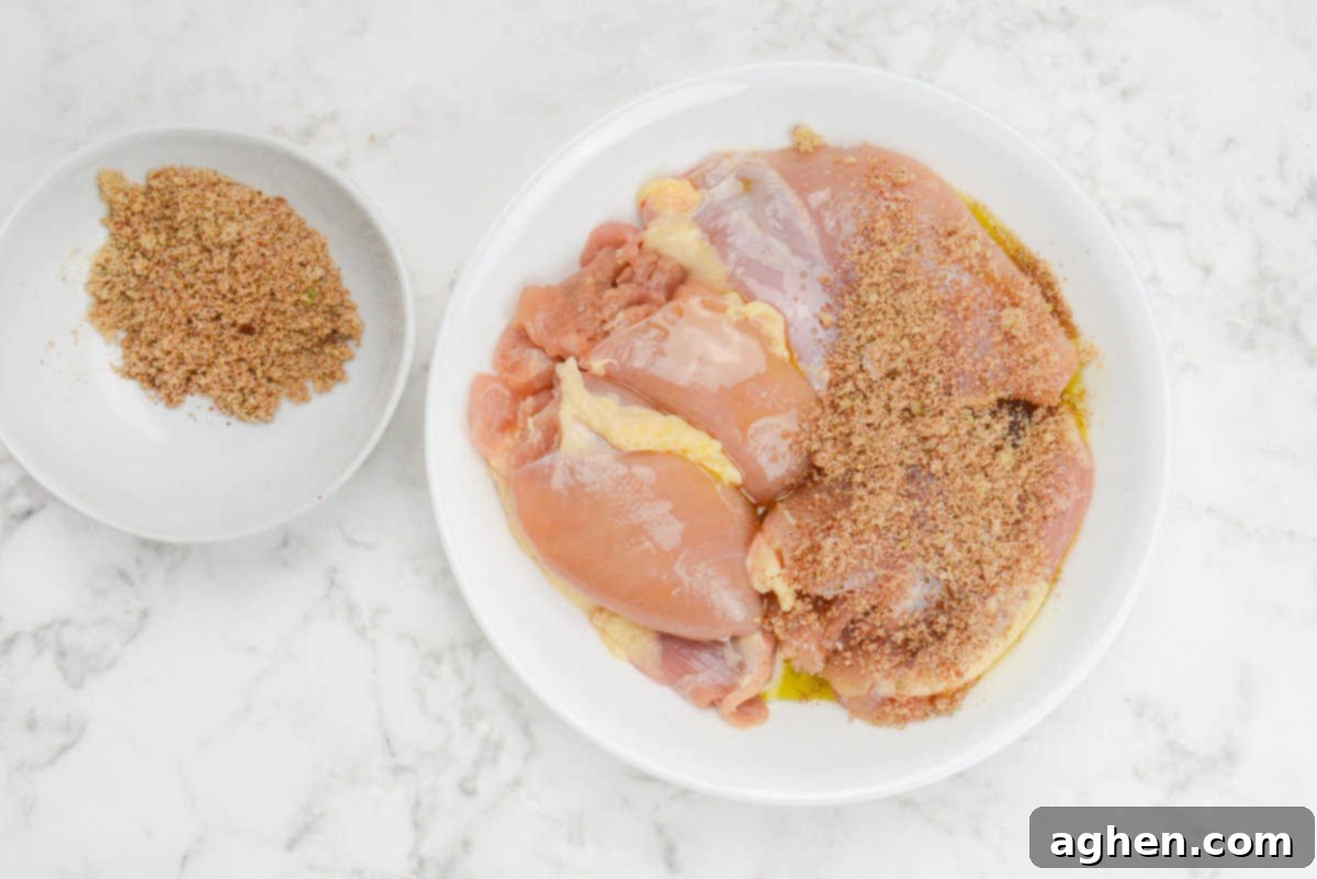 A plate of raw chicken thighs with seasoning sprinkled over half of the chicken. The remaining seasoning sits in a bowl to the side 