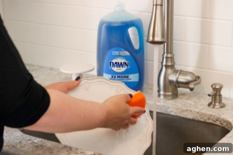 A hand washing a white plate in a kitchen sink, with a bottle of Dawn dish soap prominently displayed in the background