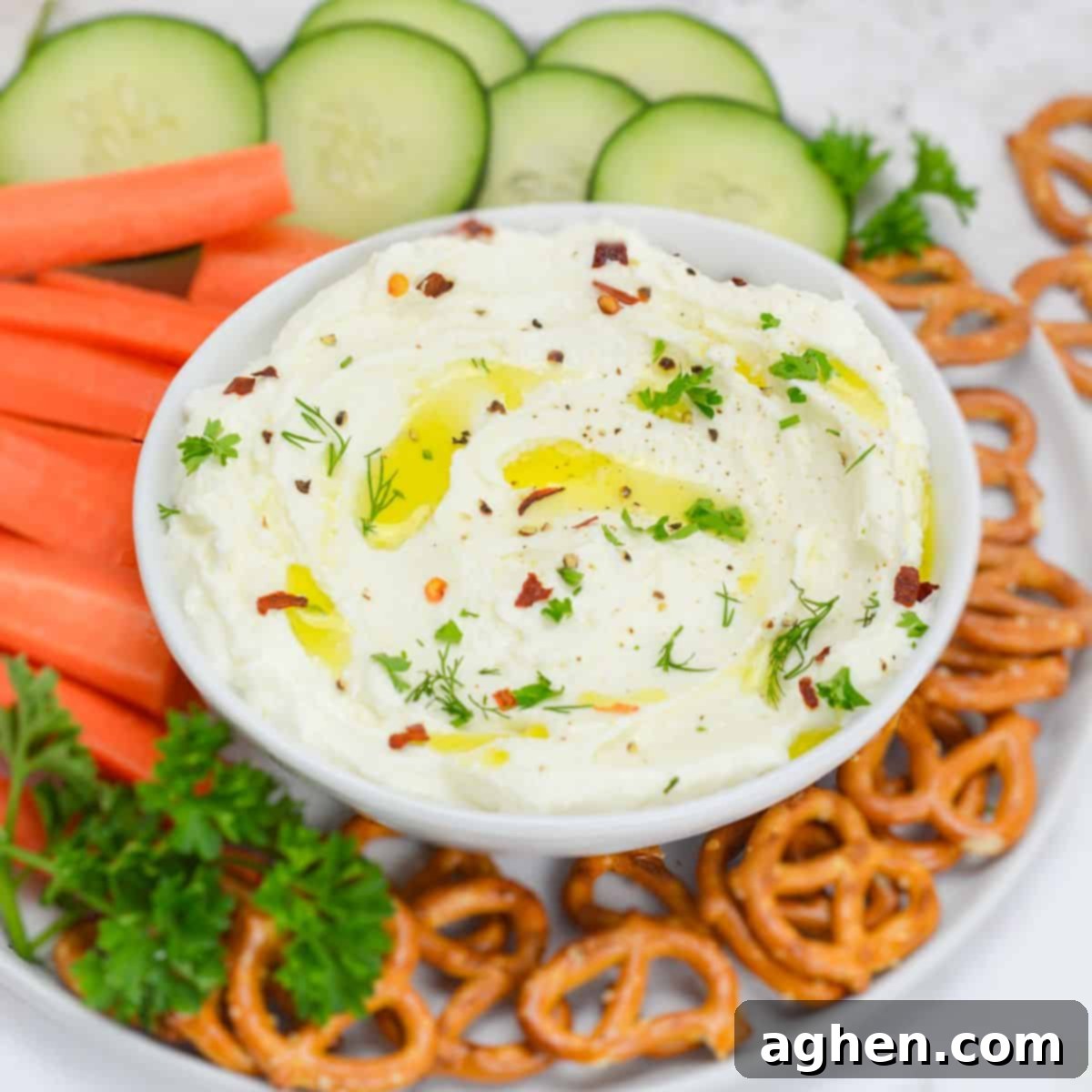Party-Perfect Weight Watchers Whipped Feta Dip 2 A close up of a white bowl filled with the finished whipped feta dip. On top of the dip are flakes of crushed red peppers, a light drizzle of oil and chopped herbs. The bowl sits on top of a plate filled with fresh cut veggies and pretzels.
