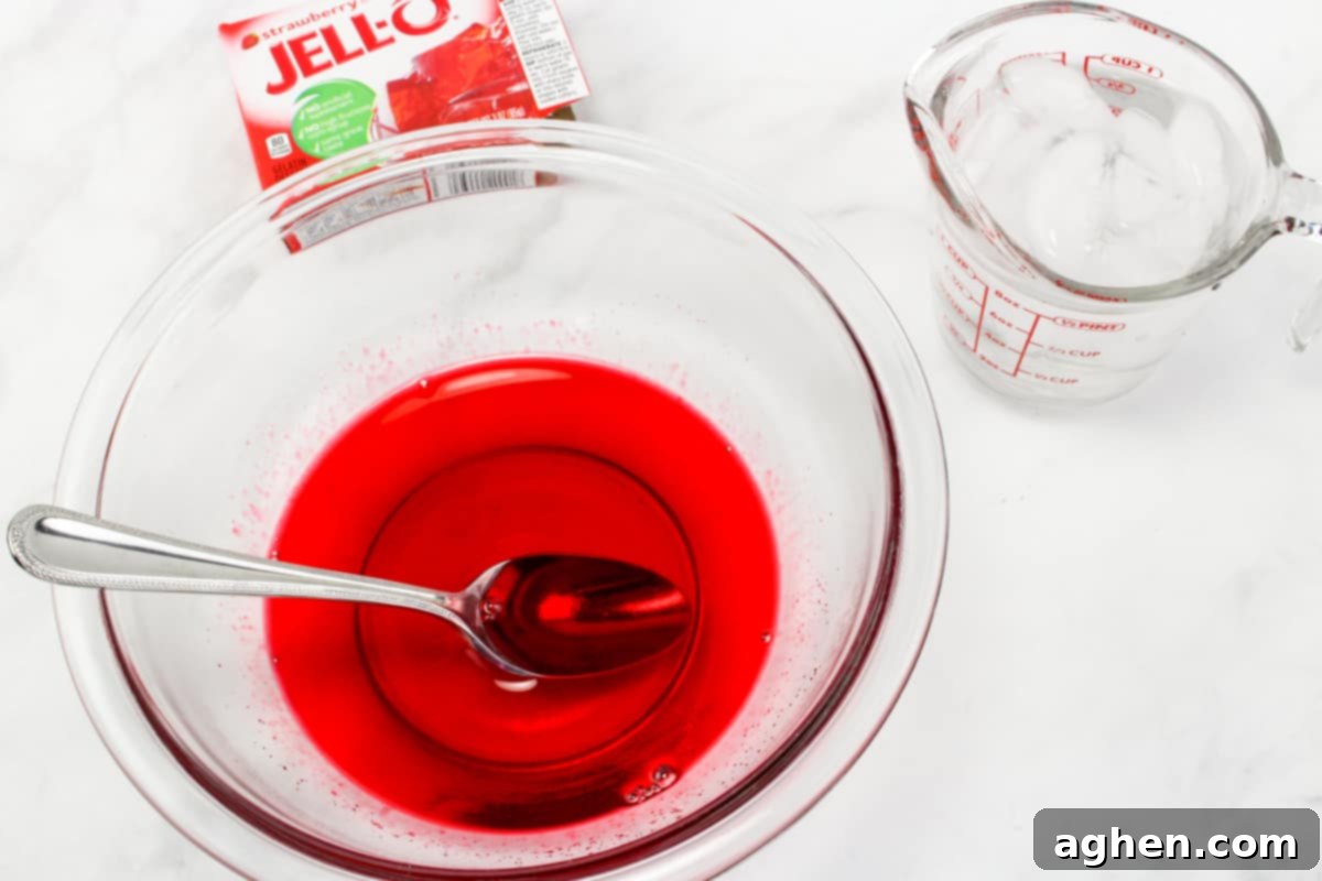 Effortless Strawberry Gem Pie 4 Bowl of jello mix dissolved in water with a spoon in it and box of strawberry jell in background.