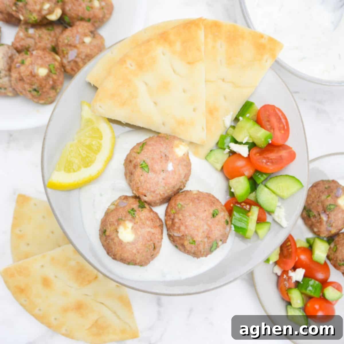 Zesty Greek Turkey Meatballs 2 An overhead view of a plate filled with baked meatballs sitting in a spoonful of tzatziki sauce. To the side is a lemon slice, pita, and a cucumber and tomato salad. In the background is an additional plate with the same ingredients, more pita and leftover meatballs.