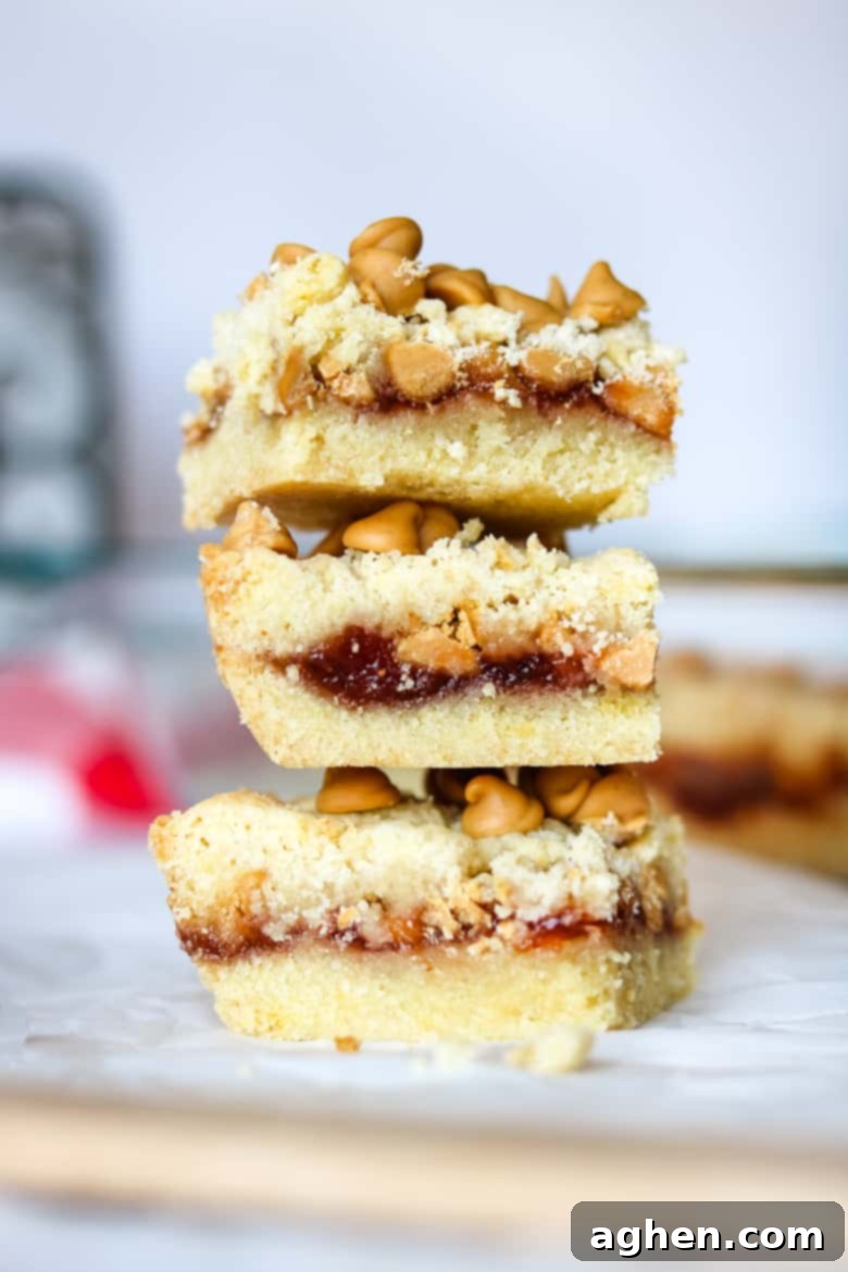 Nostalgic Peanut Butter & Jelly Squares 2 Three golden-brown peanut butter and jelly bars, generously layered with sweet fruit jam and crunchy peanut butter chips, stacked enticingly on top of one another on a white plate, ready to be enjoyed.