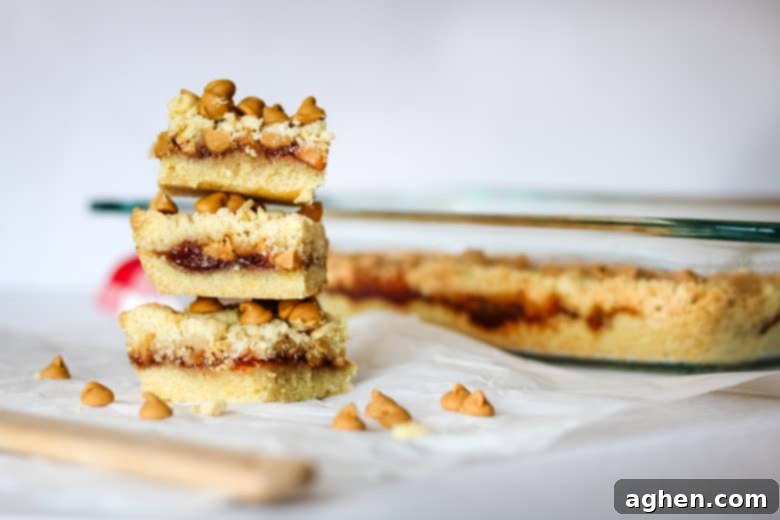 Nostalgic Peanut Butter & Jelly Squares 5 Several delicious peanut butter and jelly cookie bars, generously topped with melted peanut butter chips and a visible layer of red jam, neatly stacked on a wooden cutting board beside the glass baking pan, inviting a taste.
