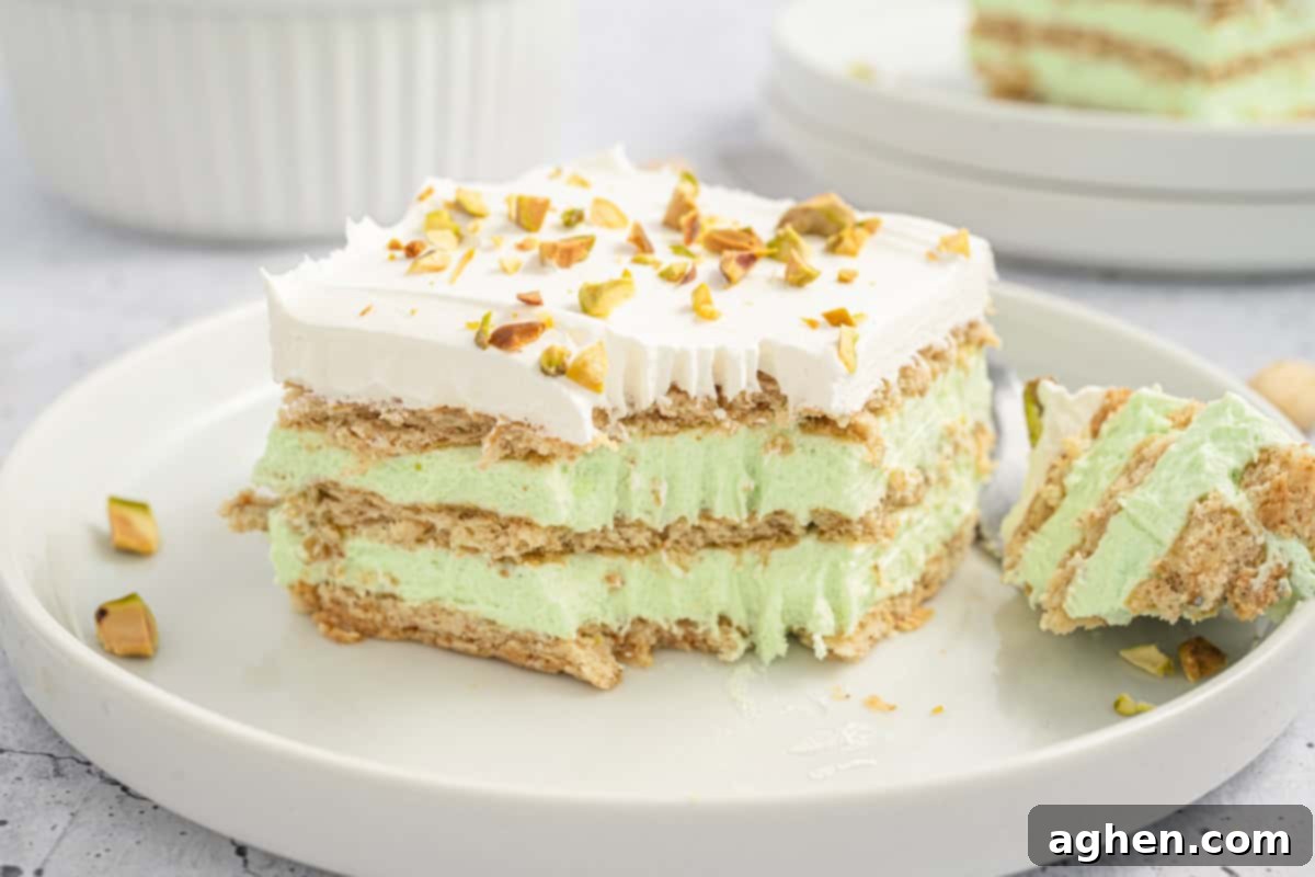 A slice of pistachio icebox cake sits on a white plate, featuring visible layers of graham crackers and pistachio-flavored filling, topped with whipped cream and crushed pistachios. A fork with a bite-sized piece rests nearby, and another plate with cake is slightly blurred in the background.