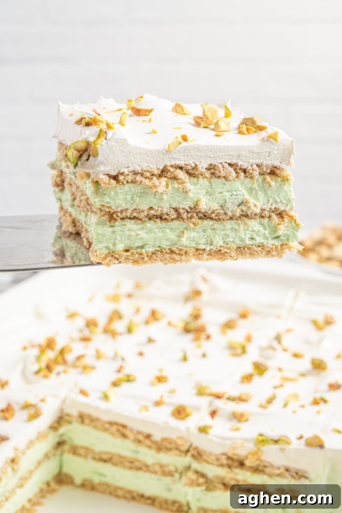 A spatula lifts a single slice of no bake pistachio icebox cake from a larger dish, showing clean layers of graham crackers, green pistachio pudding filling, and cool whip with crushed pistachios. The remaining cake is visible below.