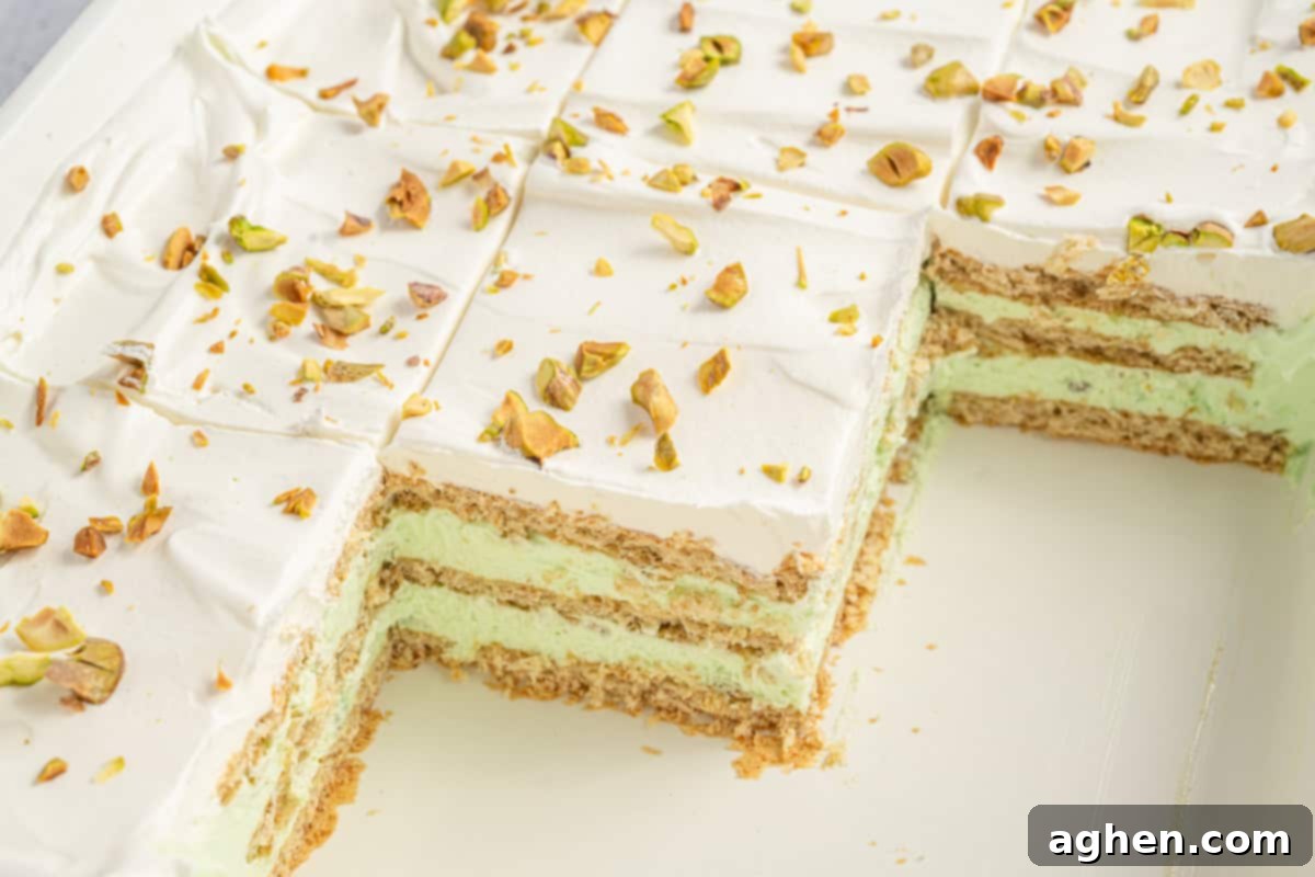 Whole pistachio icebox cake with a few slices removed, exposing the neat layers of graham crackers and green pistachio filling beneath a smooth whipped topping sprinkled with chopped pistachios.