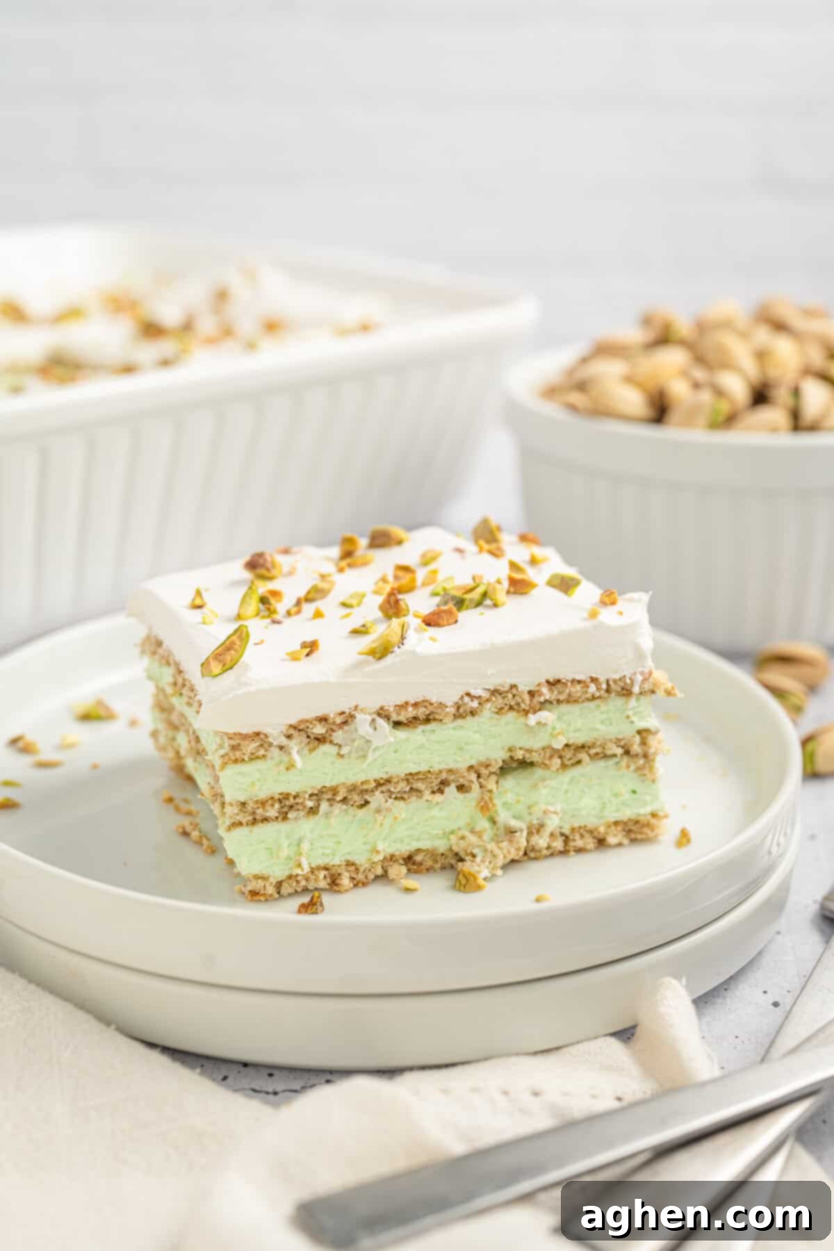 A slice of pistachio icebox cake sits on a white plate, layered with graham crackers, green pistachio filling, and whipped topping with pistachio garnish. In the background, a bowl of whole pistachios and the larger baking dish of cake are visible, creating a cozy dessert scene.