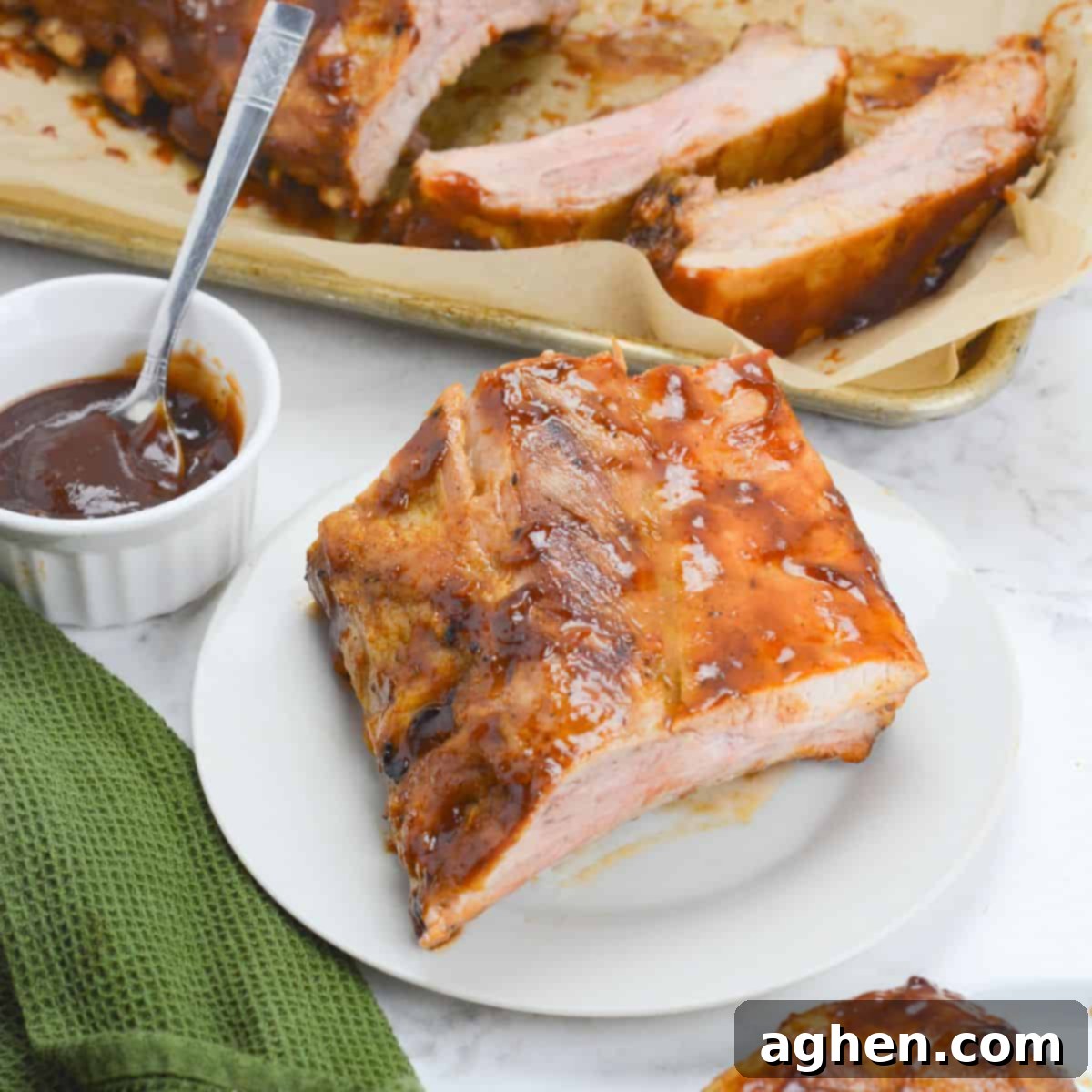 Achieving Perfect Ribs on Your Blackstone Griddle 2 A white plate with a portion of cooked ribs coated in barbecue sauce. To the side is a green textured napkin and a small bowl with sauce and a spoon. Above the plate sits a tray with sliced ribs.