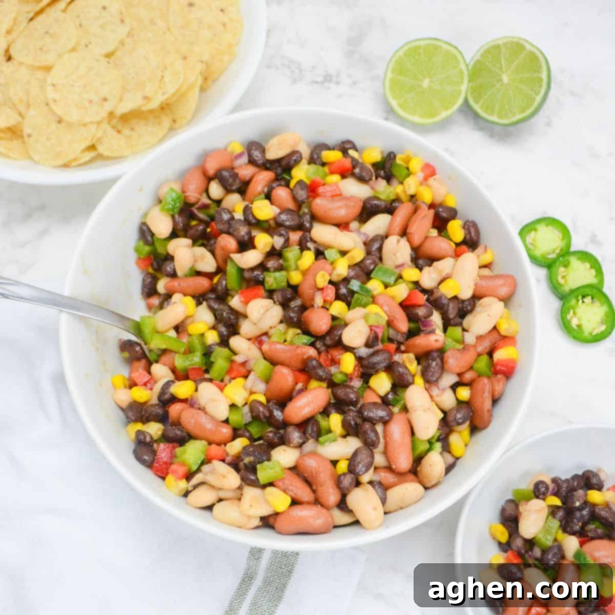 A large white serving bowl bowl filled with Mexican three bean salad. To the side is a bowl of tortilla chips, a halved lime, three slices of jalapeno and a small bowl of the salad