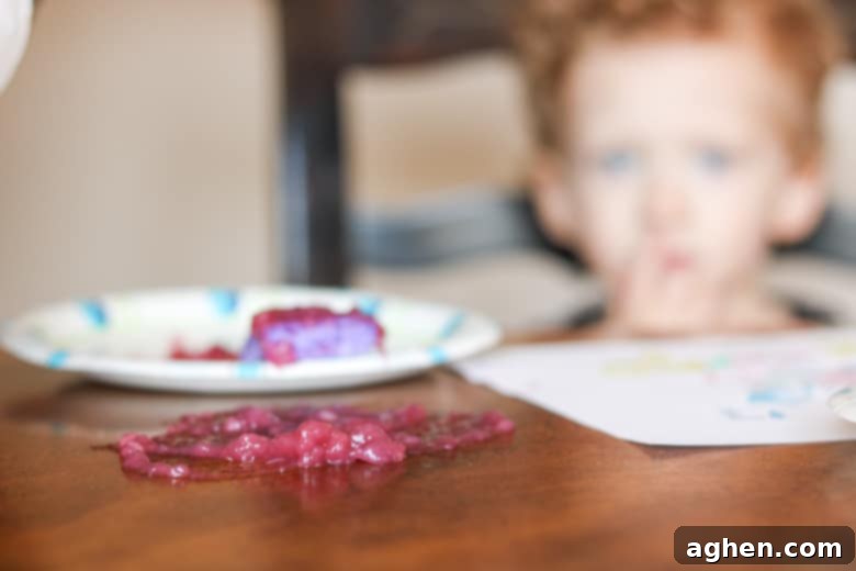 A large spill of purple paint on a dining table with a child playing in the background, illustrating a common larger mess during art time.