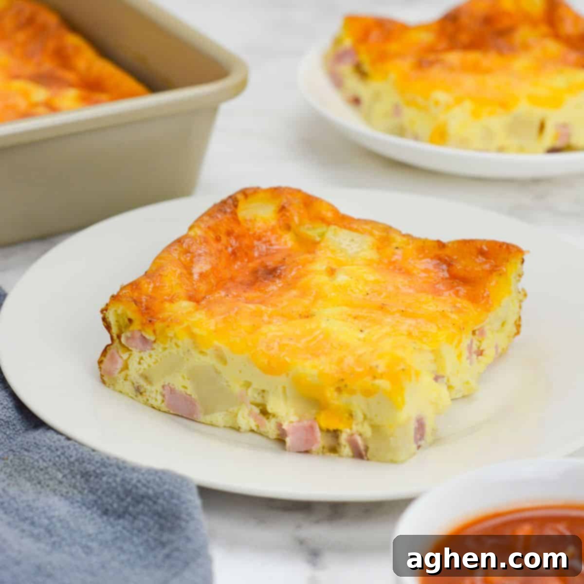 A slice of baked weight watchers breakfast casserole sits on a white plate. To the side is a small plate with a single slice, the baking pan, a gray napkin and a small bowl of salsa
