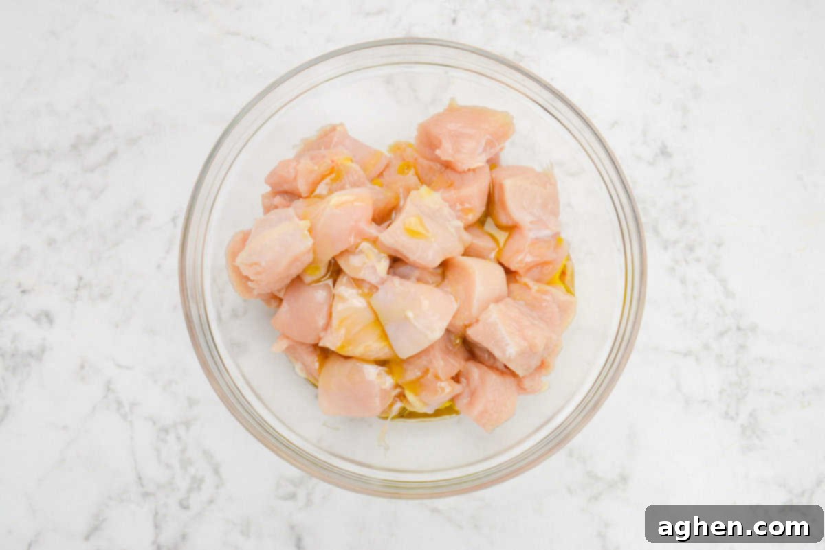 A large clear mixing bowl filled with evenly diced chicken, glistening with a yellow drizzle of olive oil, signaling the start of the marination process.