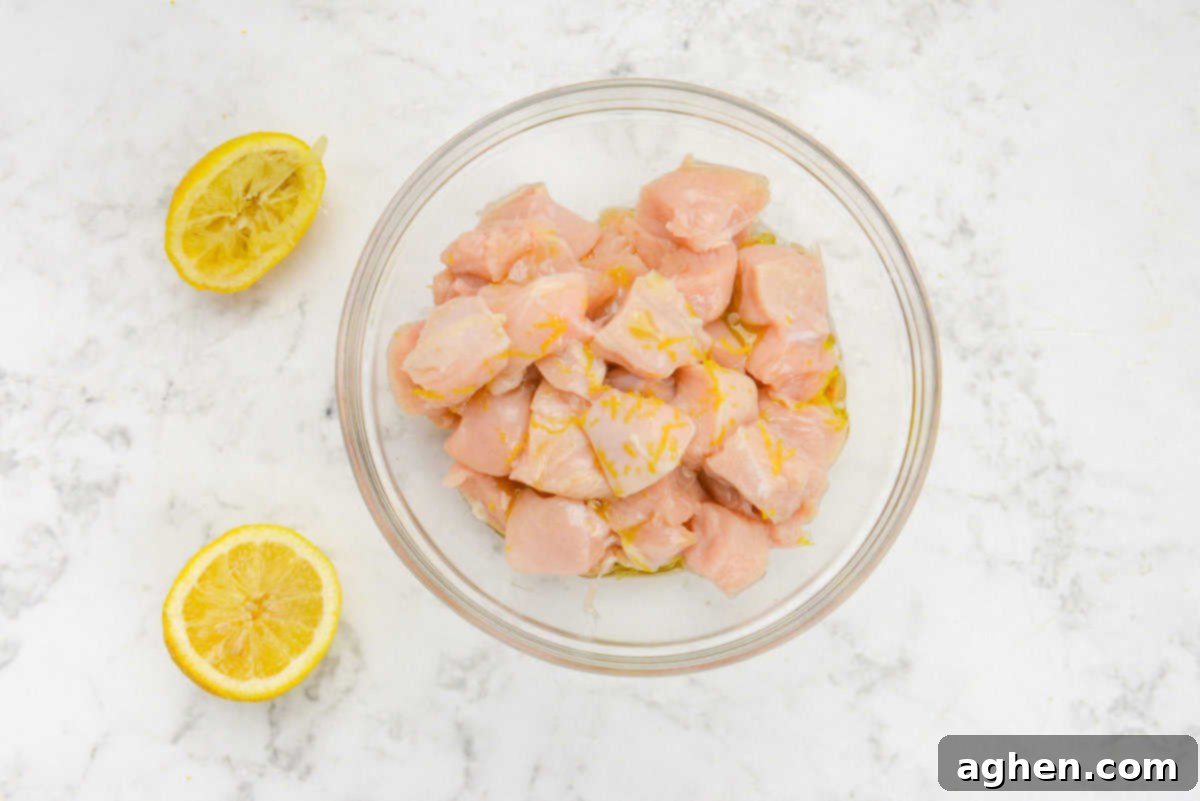 A large clear mixing bowl filled with diced chicken sits prepared, with a freshly cut lemon beside it, indicating the addition of lemon zest and juice to the marinade ingredients.