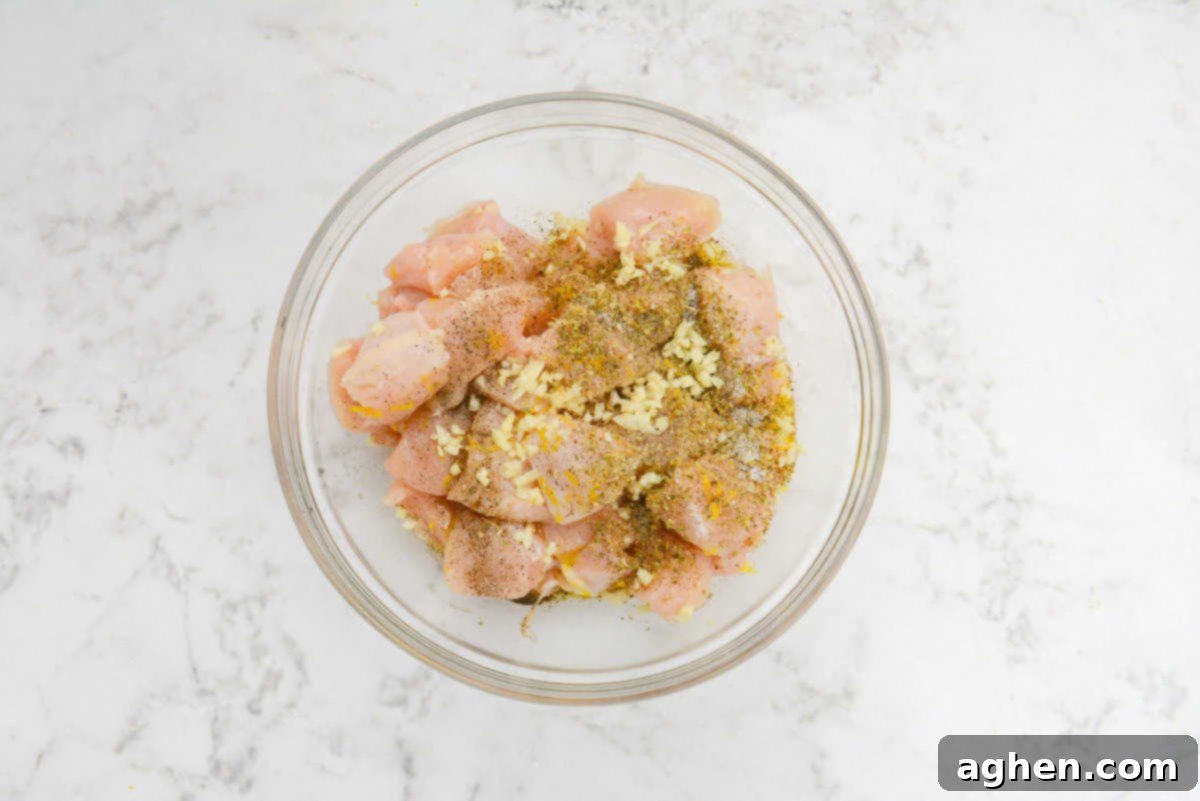 A large clear mixing bowl contains diced chicken, generously topped with an array of seasonings including minced garlic, dried oregano, salt, and pepper, all ready to be tossed and marinated for a flavorful souvlaki.