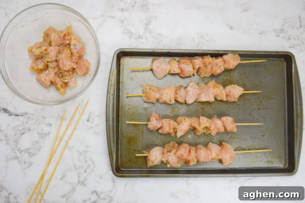 A clear mixing bowl and unused skewers are placed to the side, while in the center, a baking sheet holds four perfectly assembled chicken skewers, golden and sizzling from the oven, ready for serving.