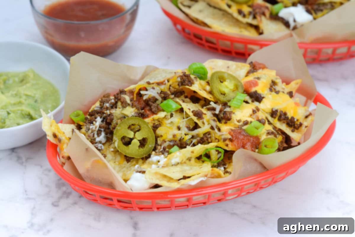 The Ultimate Blackstone Nacho Recipe 11 A red basket, lined with brown parchment paper, holds a generous serving of Blackstone nachos, beautifully topped with sliced green onions and pickled jalapenos. Small bowls of creamy guacamole and zesty salsa are positioned alongside, with another empty nacho basket ready for serving.
