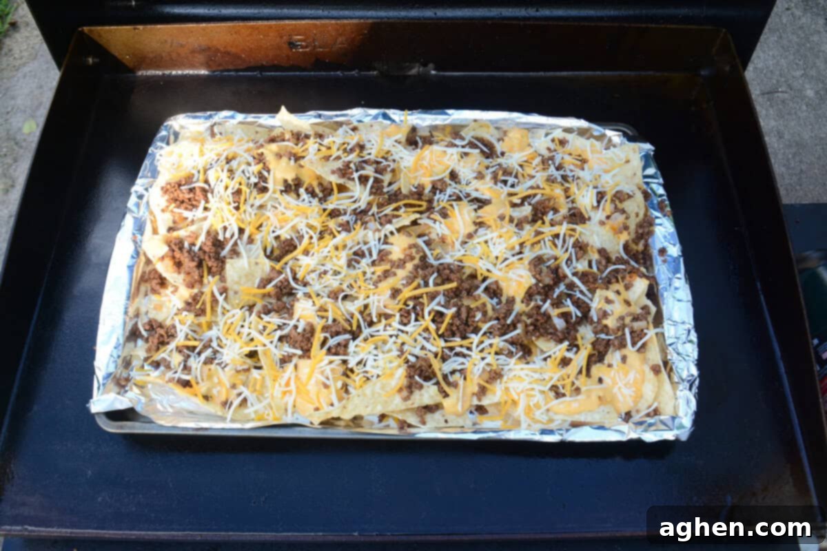 The Ultimate Blackstone Nacho Recipe 10 An overhead view of the sheet pan on the griddle, now loaded with golden tortilla chips, seasoned meat, smooth queso, and a generous topping of shredded orange and white Mexican-style cheese, ready for melting.