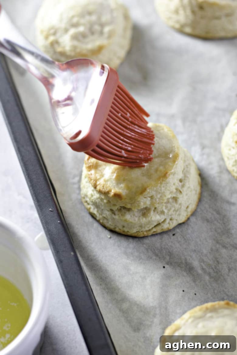 A pastry brush applying melted butter to the tops of hot, golden-brown buttermilk biscuits fresh out of the oven, enhancing their flavor and shine.
