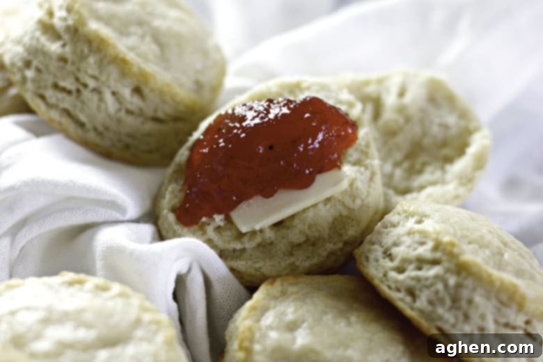 A close-up view of freshly baked buttermilk biscuits, one split open with melting butter and a dollop of bright red jam, highlighting the fluffy texture and golden crust.