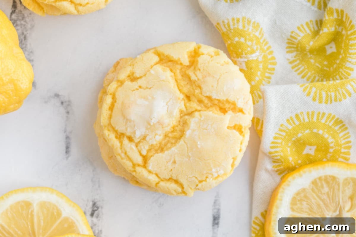 Close-up of a lemon cake mix cookie next to fresh lemons and a lemon-themed linen, highlighting its crinkled texture.