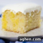 A square slice of lemon poke cake with visible Jello-infused pockets, topped with creamy frosting and a fresh lemon slice.