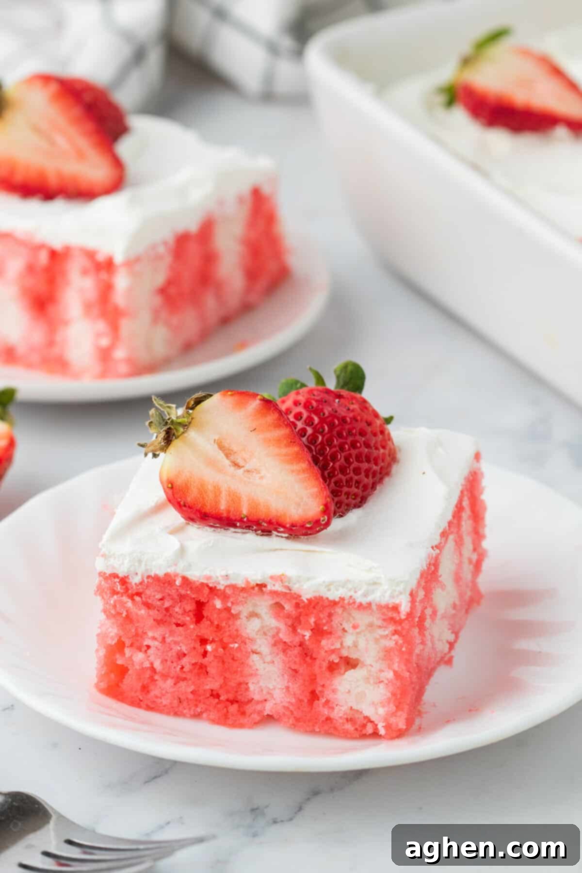 Strawberry jello poke cake topped with cool whip whipped topping and fresh strawberries and served on white plates.