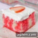 Square slice of strawberry jello poke cake made from white cake with red Jello drips through it. The cake is topped with cool whip topping and a slice of strawberry.
