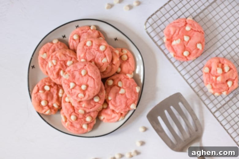 Simple Strawberry Cake Mix Cookies 5 Strawberry cake cookies with white chocolate chips on a plate, with a metal spatula and more cookies cooling on a wire rack next to it.