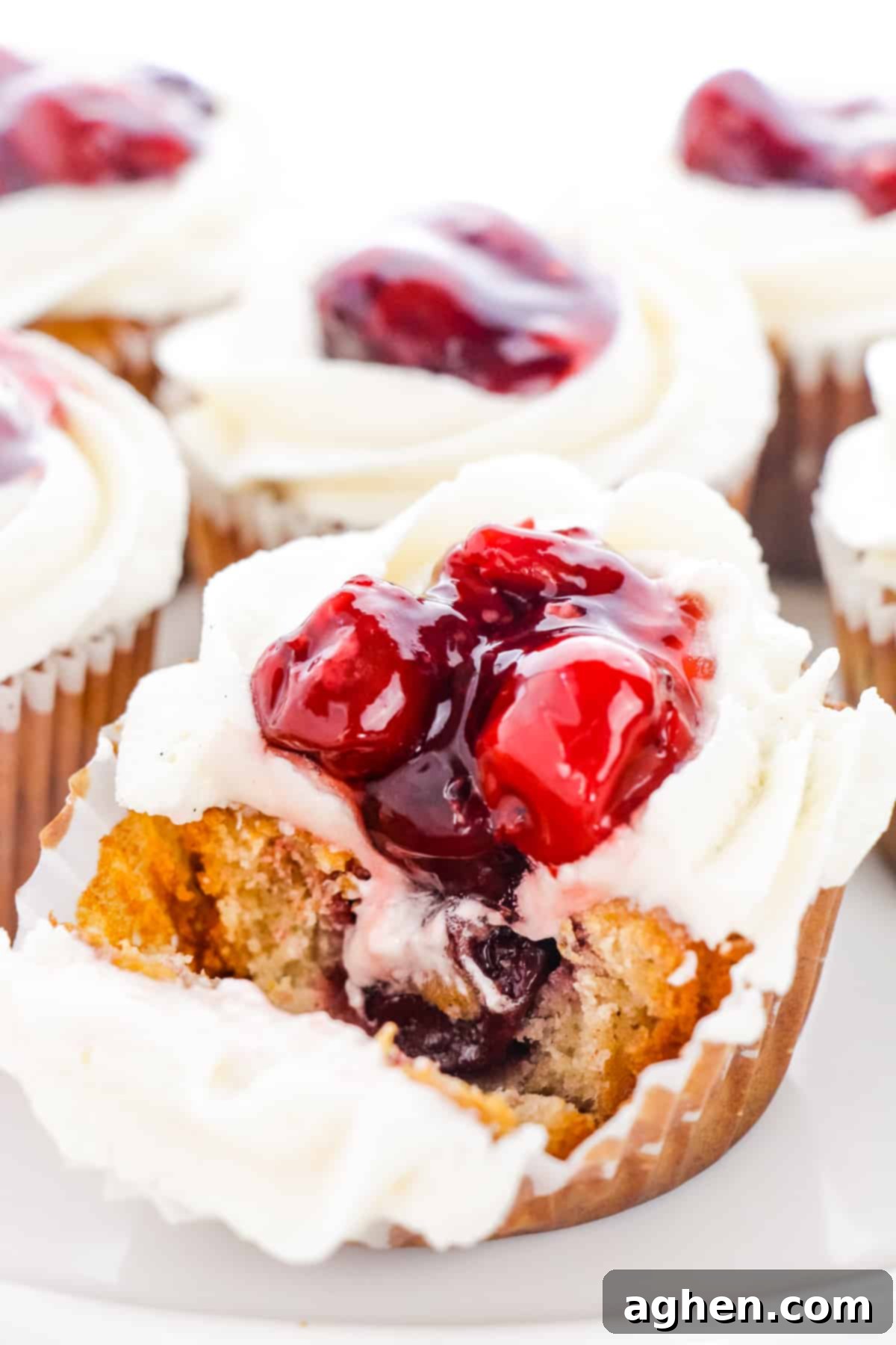 A Cherry Pie Cupcake with its liner peeled back, revealing the moist cake and the juicy cherry pie filling in the center after a bite has been taken.