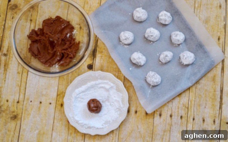 Chocolate fudge cookie dough balls being rolled generously in powdered sugar and then carefully placed on a parchment-lined baking sheet, ready for baking.