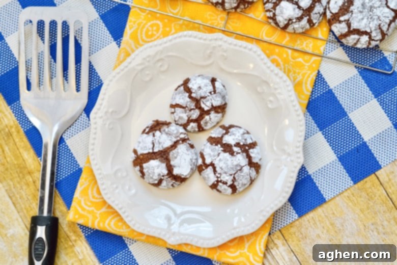 Plate of baked chocolate crinkle cookies with a spatula, with a cooling rack full of fresh cookies in the blurred background, showcasing the perfect texture and powdered sugar coating.