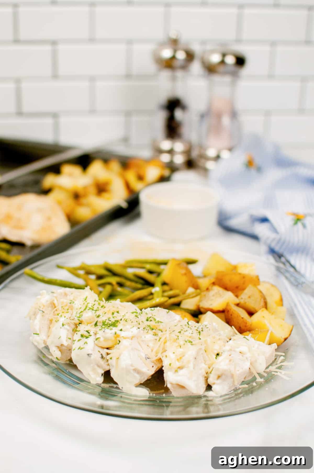 Creamy garlic herb chicken with green beans, and chopped potatoes on a clear plate with sheet pan with additional servings in background