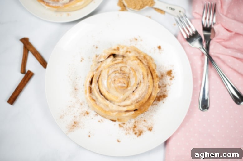 Large cinnamon roll with cream cheese frosting on a white plate with forks and cinnamon sticks next to it, air fryer dessert