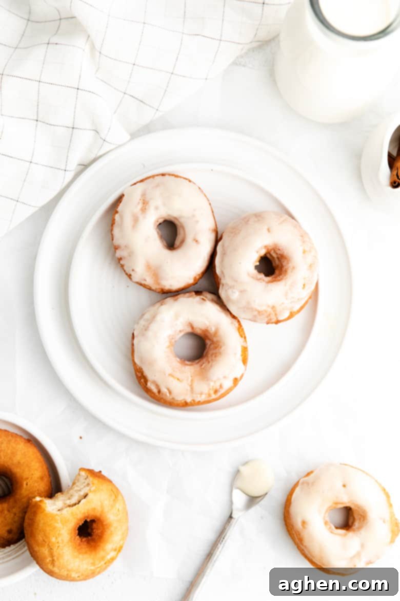 Three golden-brown glazed old-fashioned cake doughnuts stacked on a white plate, surrounded by a white linen towel, a glass jug of milk, a spoon, and additional doughnuts, some un-glazed.